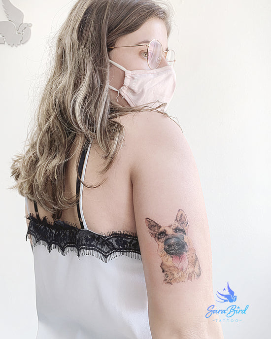 Mulher de costas a exibir a sua tatuagem na parte de trás do braço onde foi tatuado o seu cão da raça pastor alemão. A tatuagem está apenas a cabeça do cão com sua língua para fora de modo que a tatuagem ficou com o estilo realista.